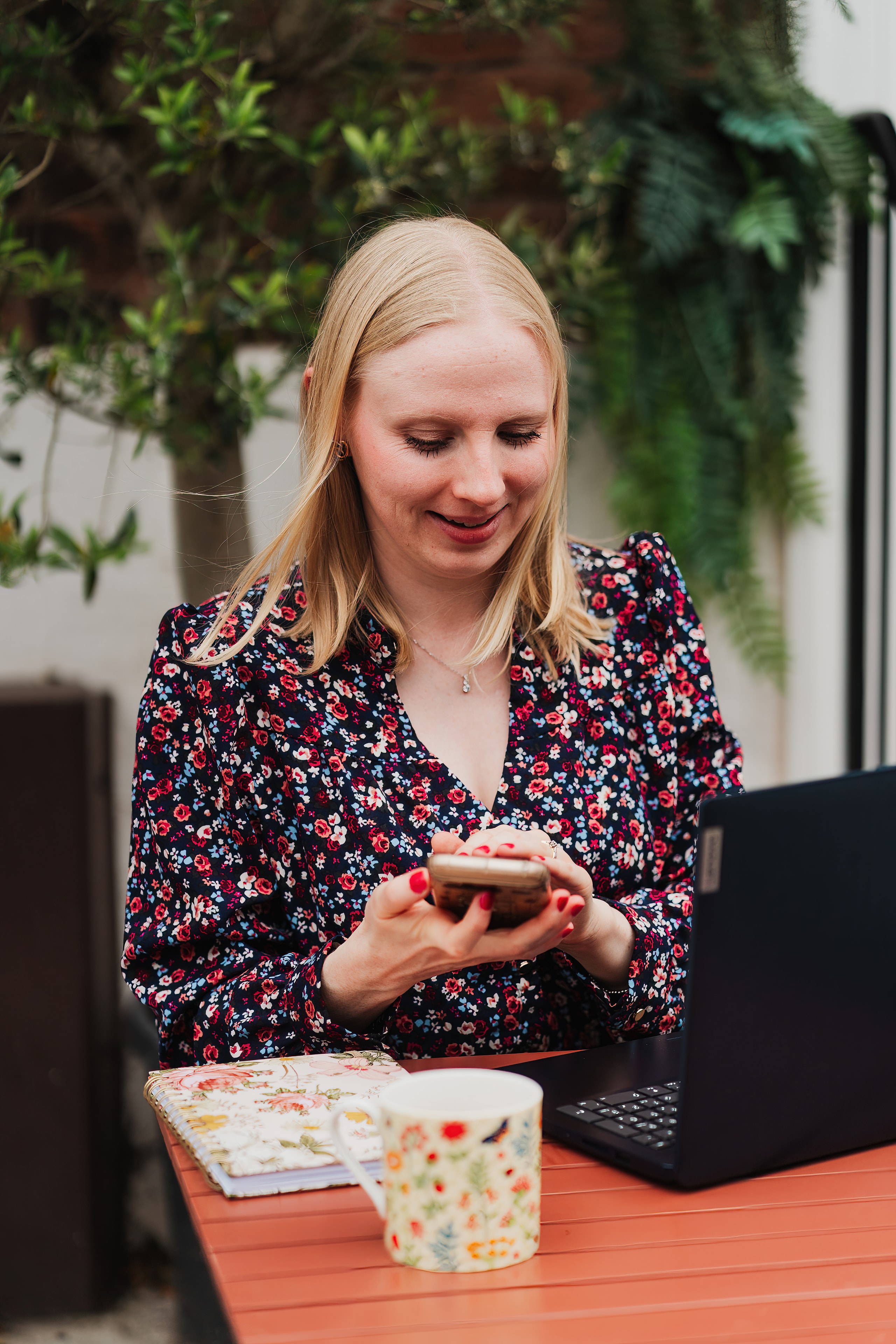 Hannah Pritchard at an outdoor table, checking her phone beside a laptop and floral mug